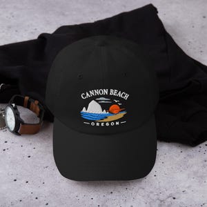 May include: A black baseball cap featuring an embroidered graphic of Cannon Beach, Oregon. The design includes a white rock formation, blue ocean waves, a tan sandy beach, and a bright orange sun, with the text CANNON BEACH OREGON in white.