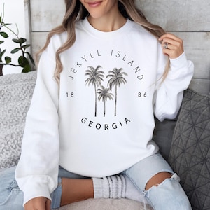 May include: White sweatshirt with black text that reads "Jekyll Island 1886 Georgia" and a graphic of three palm trees.