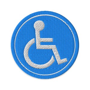 May include: A circular embroidered patch featuring a bright blue background with a white border and a white international symbol of access icon in the center, depicting a stylized figure seated in a wheelchair with a circular head shape.