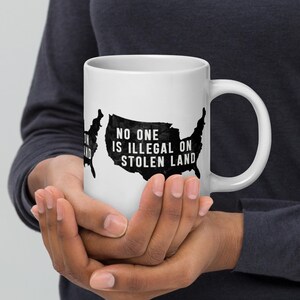 Könnte beinhalten: Eine weiße Keramiktasse mit einer schwarzen Umrandung der Vereinigten Staaten und dem Text "Niemand ist illegal auf gestohlenem Land".