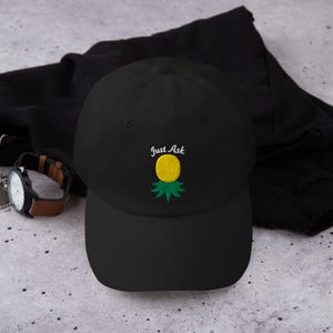 Puede incluir: Gorra de béisbol negra con un diseño de piña amarilla y el texto "Just Ask" bordado en la parte delantera. La piña tiene un cuerpo amarillo y hojas verdes. La gorra es de material suave.