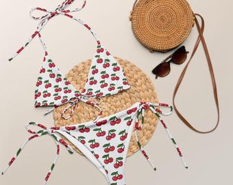 May include: White bikini set with a red cherry pattern. The bikini top has a halter neck design, and the bottoms are tie-side. A round woven bag, sunglasses, and a woven mat are also in the image.