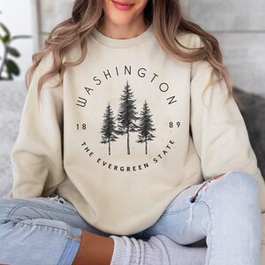 May include: A beige sweatshirt with a black graphic print of three pine trees in a circle. The text "WASHINGTON 1889 THE EVERGREEN STATE" is printed around the trees.