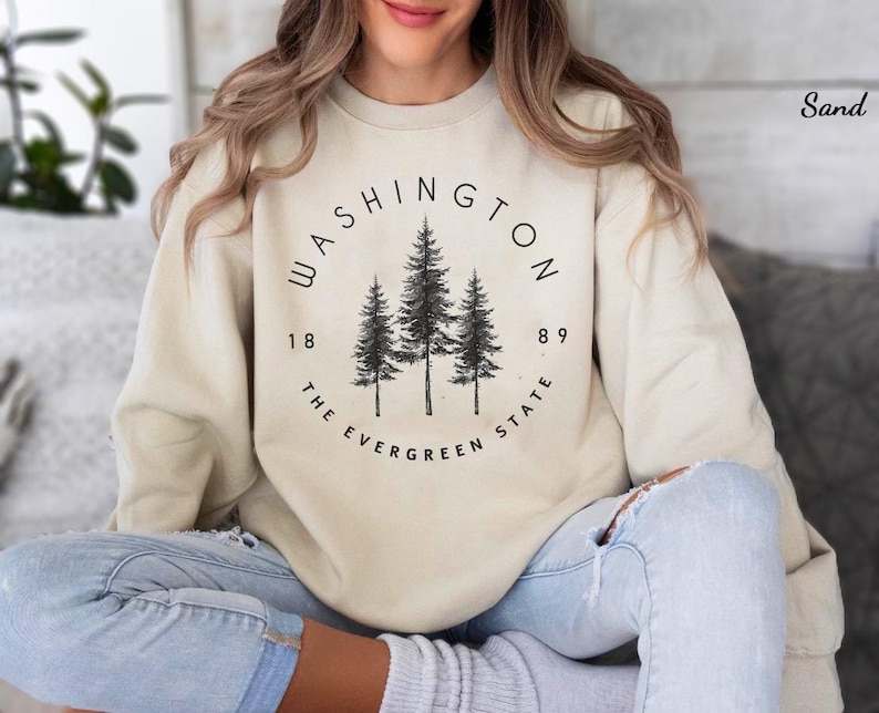May include: A beige sweatshirt with a black graphic print of three pine trees in a circle. The text "WASHINGTON 1889 THE EVERGREEN STATE" is printed around the trees.
