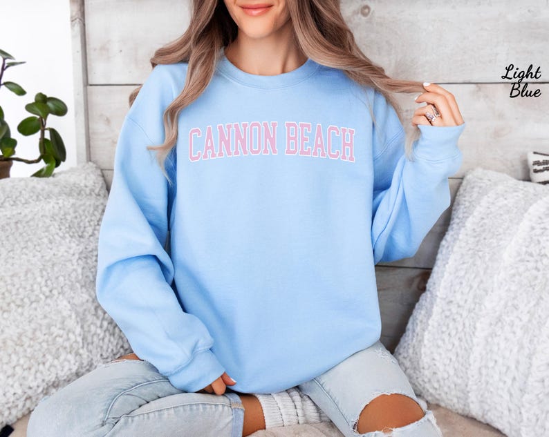 May include: Light blue sweatshirt with the text "CANNON BEACH" in pink block letters. The sweatshirt is a solid light blue color. The words "Light Blue" are written in the upper right corner.