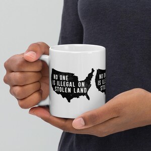 Könnte beinhalten: Eine weiße Keramiktasse mit einer schwarzen Umrandung der Vereinigten Staaten und dem Text "No one is illegal on stolen land".