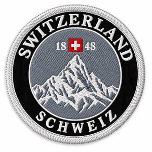 Puede incluir: Un parche bordado circular que presenta un pico de montaña en blanco y gris sobre un fondo gris. El borde es negro con el texto SWITZERLAND y SCHWEIZ en blanco. Un pequeño cuadrado rojo con una cruz blanca y 18 48 están centrados.