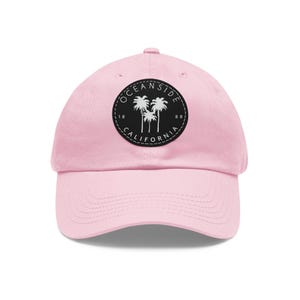May include: A light pink baseball cap with a curved brim. The cap features a circular patch with the words "Oceanside California" and palm tree silhouettes. The text and design are in black and white.