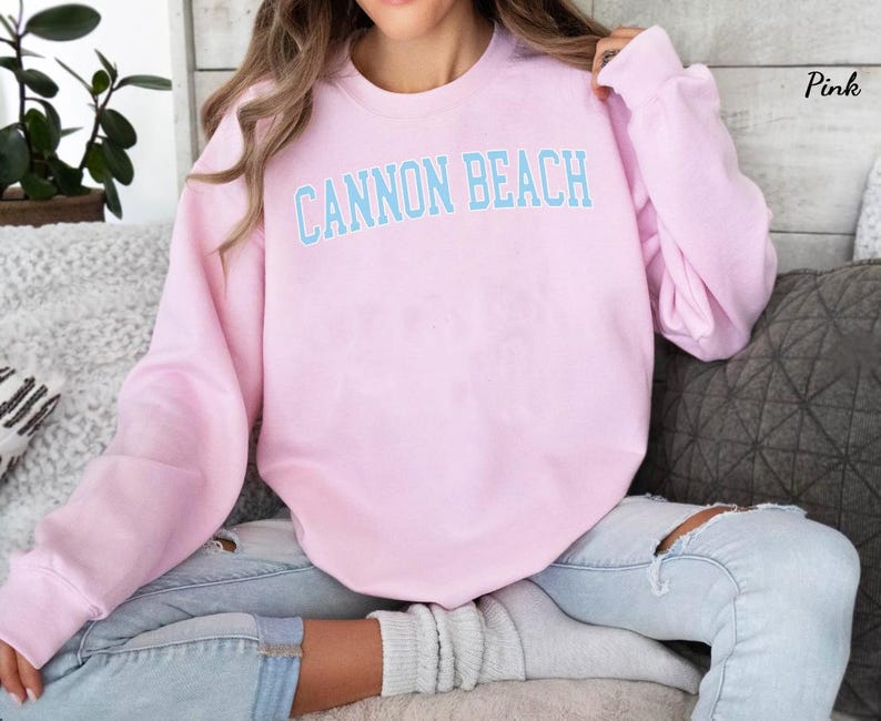May include: Pink sweatshirt with the text "CANNON BEACH" in light blue block letters. The sweatshirt is a solid pink color and has long sleeves. The text is centered on the chest.