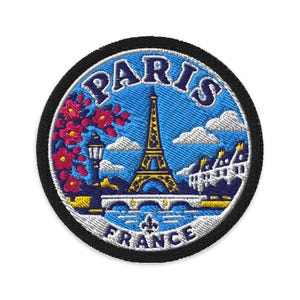 May include: A circular embroidered patch featuring a blue sky background with the Eiffel Tower in yellow and blue, a bridge, white clouds, pink flowers on the left, and the text PARIS at the top and FRANCE at the bottom in dark blue lettering.