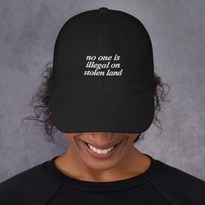 May include: Black baseball cap featuring the white text "no one is illegal on stolen land". The cap has a curved peak and is made from a soft fabric. A classic style of hat, suitable for everyday wear.
