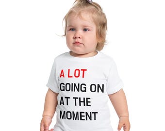 Op de afbeelding: Een wit t-shirt met de tekst "A LOT GOING ON AT THE MOMENT" in rood en zwart. Het shirt wordt gedragen door een jong kind met blond haar.