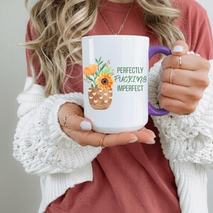 May include: White ceramic mug with a purple handle, featuring a floral design with sunflowers and the text "Perfectly Fucking Imperfect" in green. The mug has a brown vase design with heart-shaped cutouts.