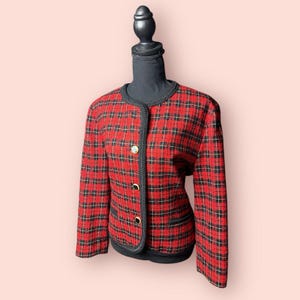 May include: A red and black plaid jacket with a black trim and gold buttons. The jacket is displayed on a black mannequin. The jacket has a classic, tailored design, with a round neckline and long sleeves. The plaid pattern is a bold, eye-catching design.