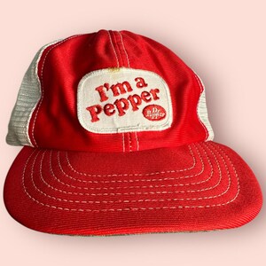 Pode incluir: Boné de beisebol vermelho e branco com o texto bordado "I'm a Pepper" em um patch. O boné apresenta uma aba curva e uma parte traseira de malha. O logotipo da Dr. Pepper também está no patch.