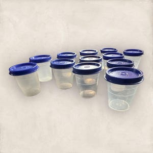 Puede incluir: Un conjunto de vasos de plástico transparente con tapas azul oscuro. Los vasos están dispuestos en grupo, algunos ligeramente inclinados. Las tapas tienen un diseño circular. Los vasos parecen estar vacíos.