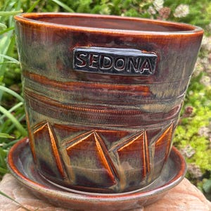 May include: A cylindrical ceramic planter with a matching saucer, featuring a glossy glaze in shades of brown, copper, and green. The exterior displays a horizontal band of textured lines above carved triangular shapes and the word SEDONA.