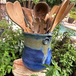 May include: A collection of wooden kitchen utensils, including spoons and spatulas, displayed in a blue and tan ceramic utensil holder. The holder is surrounded by green plants and sits on a rock.