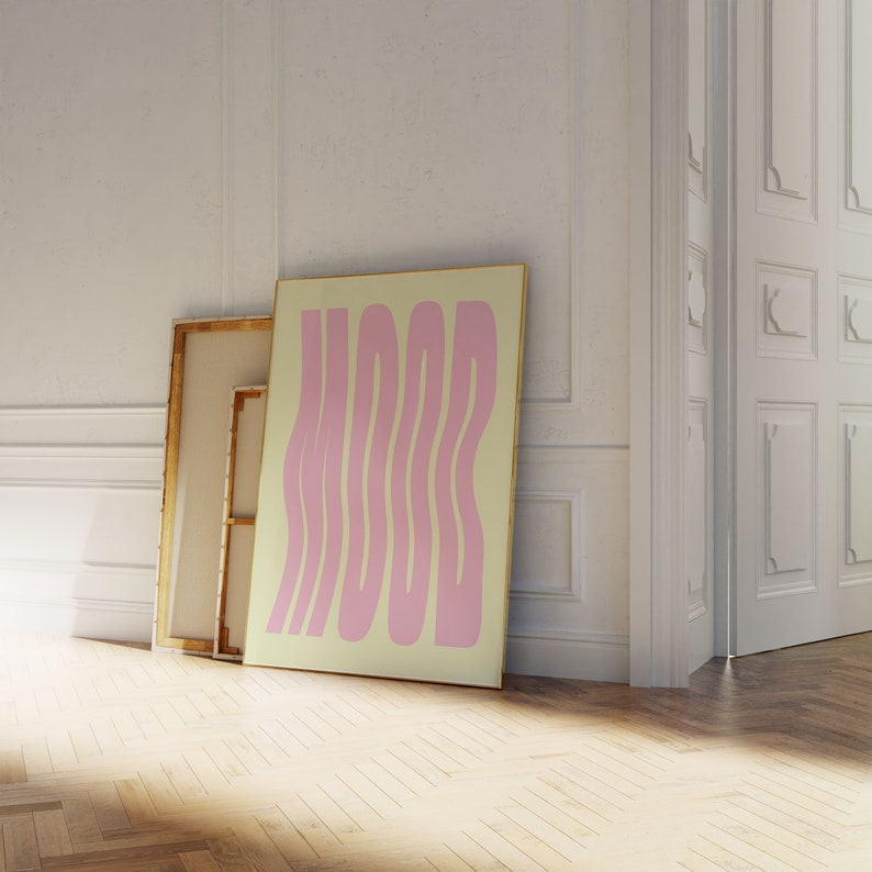 May include: A framed print with a yellow background and pink wavy text that reads "MOOD". The print is leaning against a white wall next to a wooden door.