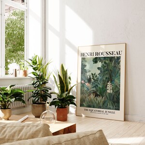 May include: A framed poster of a painting by Henri Rousseau titled "The Equatorial Jungle" featuring a lush green jungle scene with a tiger in the foreground. The poster is hanging on a white wall in a room with plants and a wooden coffee table.