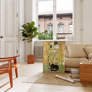 May include: A framed print of Gustav Klimt's "The Kiss" with a black cat lying at the bottom. The artwork features gold, yellow, and black colors. The print is displayed in a bright, airy room with a beige sofa and wooden furniture.