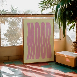 May include: A framed print with a yellow background and pink wavy text that reads "MOOD". The print is leaning against a wall with a window behind it.