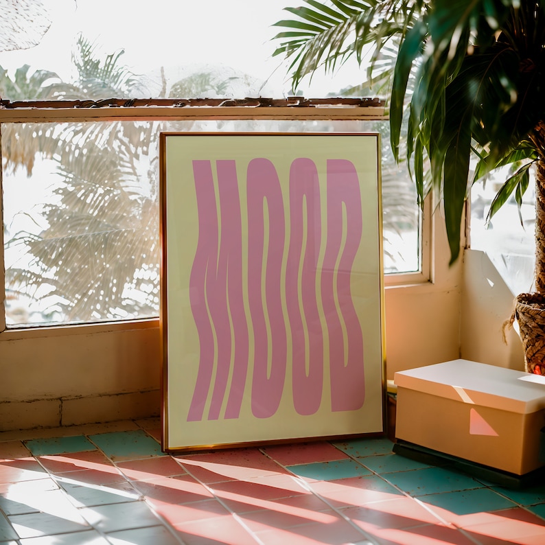 May include: A framed print with a yellow background and pink wavy text that reads "MOOD". The print is leaning against a wall with a window behind it.