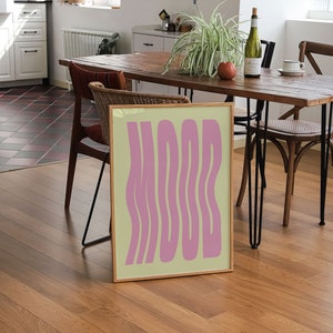 May include: A framed print with a light yellow background and the word "MOOD" in pink, wavy letters. The print is sitting on a wooden table in a dining room.