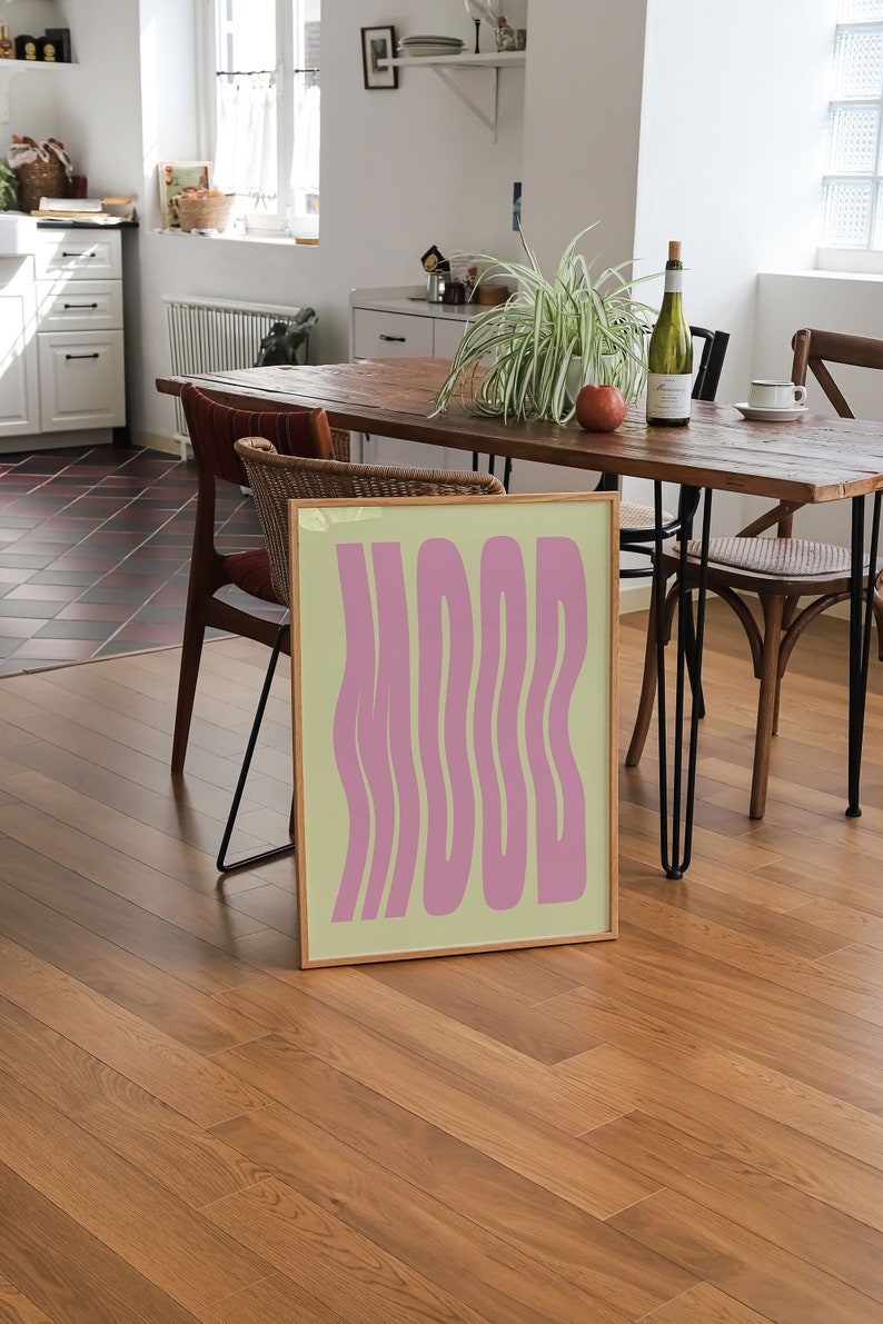 May include: A framed print with a light yellow background and the word "MOOD" in pink, wavy letters. The print is sitting on a wooden table in a dining room.