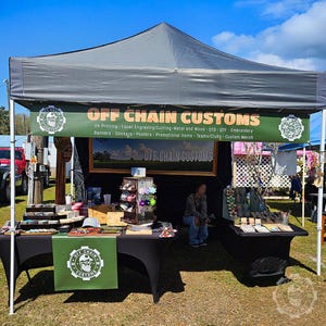 May include: A black and green banner with the text "OFF CHAIN CUSTOMS" and a list of services offered. The banner is hanging from a black canopy tent. There is a table with various items for sale, including stickers, posters, and promotional items. The table is covered with a black tablecloth. There is a person sitting in the background.