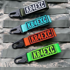 May include: Four nylon webbing keychains with black plastic carabiner clips. Each strap features a rectangular embroidered patch with the text KR4EXC. The straps are olive green, turquoise, orange, and neon green with matching or black lettering.