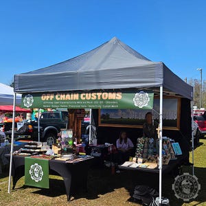 May include: A black canopy tent with a sign that reads "OFF CHAIN CUSTOMS" and offers UV printing, laser engraving, cutting, metal and wood, OTG, OTF, embroidery, banners, stickers, posters, promotional items, teams/clubs, and custom merch. The tent is set up at an outdoor market with tables and chairs.