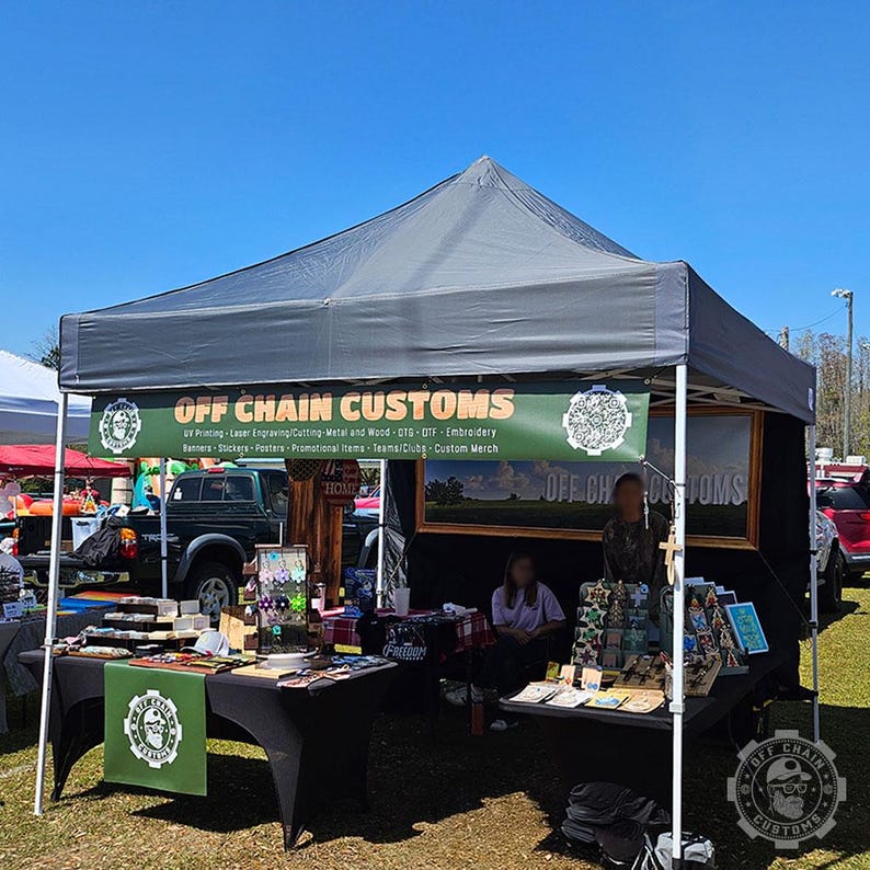 May include: A black canopy tent with a sign that reads "OFF CHAIN CUSTOMS" and offers UV printing, laser engraving, cutting, metal and wood, OTG, OTF, embroidery, banners, stickers, posters, promotional items, teams/clubs, and custom merch. The tent is set up at an outdoor market with tables and chairs.
