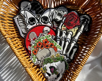 May include: A collection of embroidered iron-on patches featuring various skull designs, including a skull with flowers, a skull with a red background, and a skeleton hand holding a rose. The patches are in a wicker heart-shaped basket.
