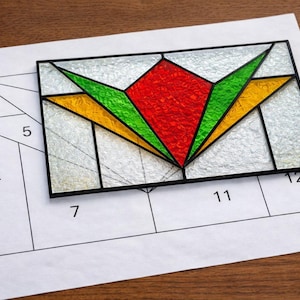 May include: A stained glass panel with a geometric design in red, green, and yellow, set against a clear background. The panel is placed on a paper pattern with numbered sections, likely for a craft project. The panel is rectangular with black borders.