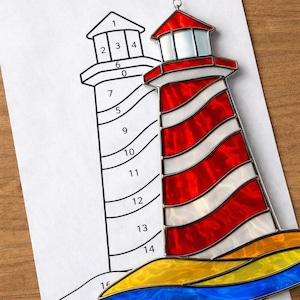 May include: A stained glass lighthouse with red and white stripes, a red roof, and a blue and yellow base. A black and white line drawing of the lighthouse is next to the stained glass version, with numbers for color placement.