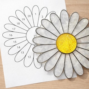 Peut inclure: Une marguerite en vitrail avec des pétales blancs et un centre jaune, à côté d'un motif numéroté sur papier. La marguerite a un contour noir et est sur une surface en bois.