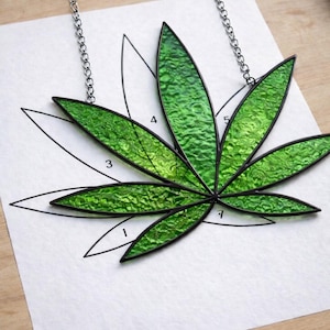 May include: A green stained glass leaf pendant with a silver chain. The leaf design has a textured surface and black outlines. The pendant is resting on a white paper with a leaf outline and numbers.