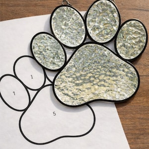 May include: A stained glass paw print with a textured, clear glass design and black borders. The paw print is placed on top of a paper template with a black outline and numbers. The background is a wooden surface.