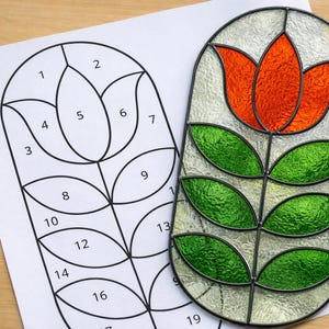 May include: A stained glass panel in an oval shape, featuring a vibrant orange tulip flower with green leaves and a stem. A black and white line drawing of the same flower is next to it, with numbered sections for a craft project.