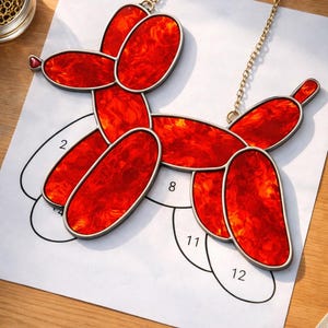 May include: A red stained glass balloon dog with a gold chain for hanging. The dog is made of red glass with a textured appearance and a silver outline. The dog is resting on a white paper with numbers.