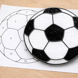 May include: A black and white soccer ball made of glass, resting on a white paper with a soccer ball outline. The ball has black pentagons and white hexagons. The paper has numbers inside the outline.