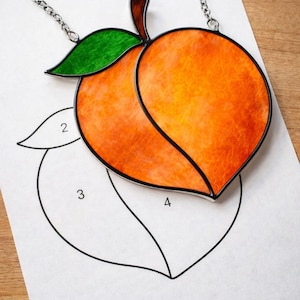 May include: A stained glass peach pendant with a silver chain. The peach is orange with a green leaf and brown stem. The pendant is on a white paper with a peach outline.