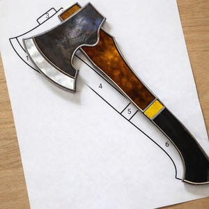 May include: An axe with a silver blade and a brown and black handle. The axe head has a dark, textured appearance. The handle features a yellow accent near the center and a black base. The axe is on a white paper with a black outline.