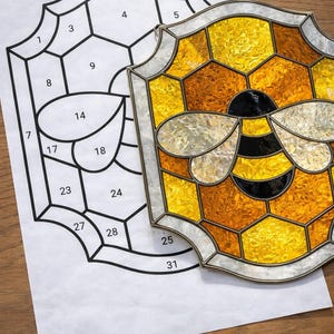Puede incluir: Una abeja de vidriera con detalles amarillos, naranjas, negros y plateados, apoyada sobre un patrón numerado. El diseño de la abeja presenta un patrón de panal y está enmarcado con un borde plateado. El patrón está sobre una hoja de papel blanca.