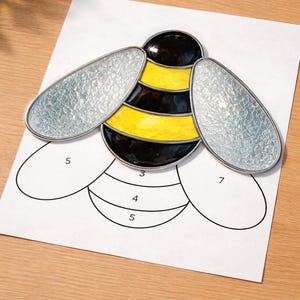 May include: A stained glass bee craft project with a black and yellow body, silver wings, and a black outline. The bee is placed on a white paper with numbered sections, ready for assembly. The project is on a wooden surface.