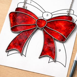 May include: A red stained glass bow pendant with a silver chain. The bow is a vibrant red with a textured appearance, outlined in silver. It is placed on a white paper with a bow outline and numbers. A pen is visible on the right.