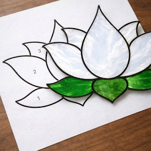 May include: A stained glass lotus flower design with white petals and green leaves, outlined in black. The design is on a white paper with numbers 1, 2, and 3 labeling the petals. The background is a wooden surface.