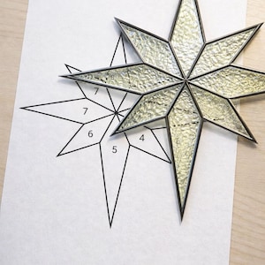 May include: A clear, textured glass star with a dark metal frame, resting on a white paper with a star pattern and numbers. The star has a loop for hanging. The background is a light wood surface.