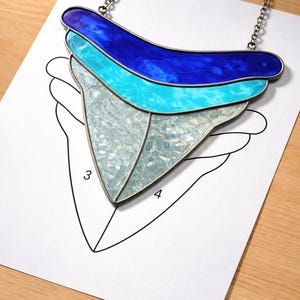 May include: A stained glass necklace in shades of blue and silver, resembling a shark tooth. The pendant hangs from a silver chain and rests on a paper template with numbers 3 and 4.